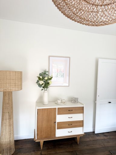 Buffet en bois clair avec vase de fleurs, lampe en osier, et peinture au mur.